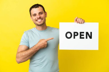 Mor arkaplan üzerinde Brezilyalı bir adam açık yazılı bir pankart tutuyor ve onu işaret ediyor.