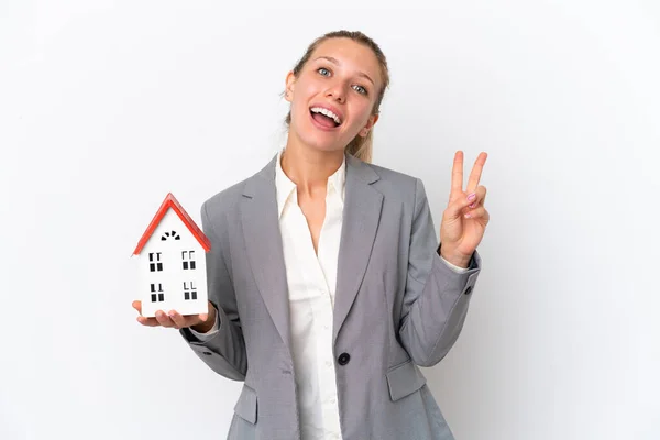 房地产商女人抱着一间因白色背景而被隔离的玩具屋 笑着表示胜利的信号 — 图库照片