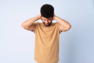 Faslı genç bir adam mavi arka planda izole edilmiş hayal kırıklığına uğramış ve kulaklarını kapatmış.