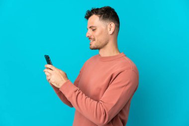 Mavi arka planda izole edilmiş genç beyaz adam cep telefonuyla mesaj ya da e-posta gönderiyor.