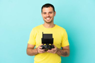 Brezilyalı adam uzaktan kumandayla izole edilmiş mavi arkaplanı kontrol ediyor. Kolları kalçasında ve gülümsüyor.