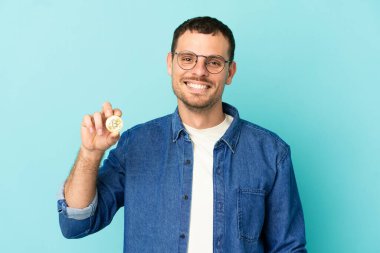 Elinde Bitcoin tutan Brezilyalı bir adam izole edilmiş mavi arka planda kolları kalçasında ve gülümsüyor.