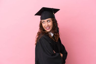 Üniversiteden mezun genç bir kadın pembe arka planda izole edilmiş, kolları bağlı ve mutlu.