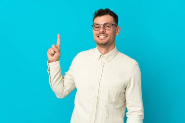 Beyaz tenli yakışıklı adam mavi arka planda izole edilmiş ve en iyinin işareti olarak parmağını kaldırıyor.