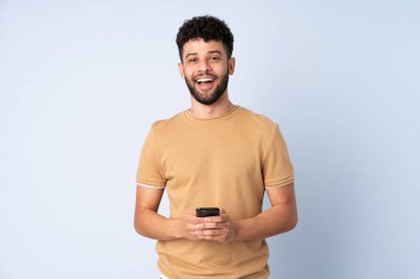 Faslı genç bir adam mavi arka planda izole edilmiş, şaşırmış ve bir mesaj gönderiyor.