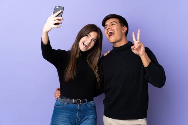 İzole edilmiş mor arka planı olan genç bir çift cep telefonuyla selfie çekiyor.