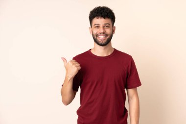 Bej arka planda izole edilmiş genç Faslı bir adam bir ürün sunmak için kenarı işaret ediyor.