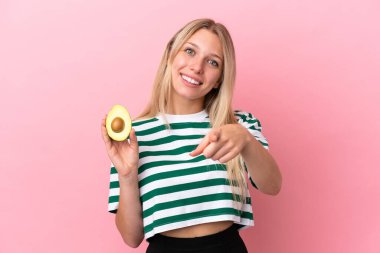 Avokado tutan beyaz bir kadın pembe arka planda izole edilmiş mutlu bir ifadeyle ön tarafı işaret ediyor.