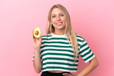 Avokado tutan beyaz bir kadın pembe arka planda izole edilmiş. Kolları kalçasında ve gülümsüyor.
