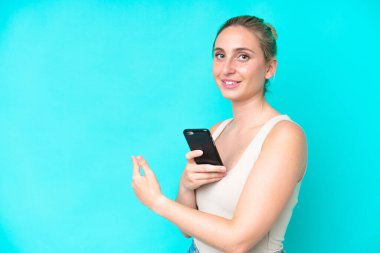 Genç beyaz kadın mavi arka planda izole edilmiş cep telefonu kullanıyor ve geri işaret ediyor.