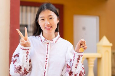 Genç Çinli bir kadın evin anahtarlarını tutuyor. Gülümsüyor ve zafer işareti gösteriyor.