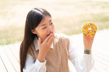 Genç Çinli bir kadın sürpriz ve şok olmuş bir yüz ifadesiyle dışarıda donut tutuyor.