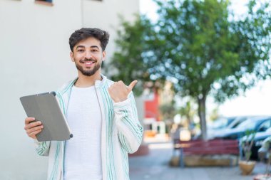 Dışarıda elinde tablet tutan yakışıklı bir Arap bir ürünü sunmak için kenarı işaret ediyor.