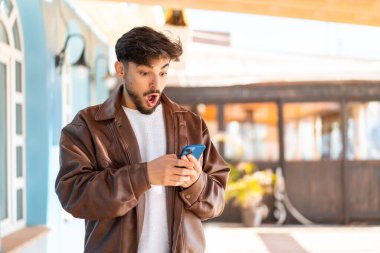 Dışarıdaki yakışıklı Arap adam şaşırmış bir ifadeyle cep telefonunu kullanırken kameraya bakıyor.