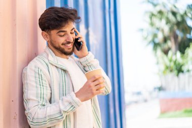 Dışarıdaki yakışıklı Arap adam cep telefonu kullanıyor ve elinde mutlu bir ifadeyle kahve tutuyor.