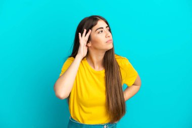 Mavi arka planda izole edilmiş genç beyaz kadın elini kulağına koyarak bir şeyler dinliyor.