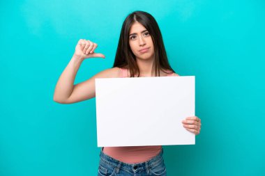 Young caucasian woman isolated on blue background holding an empty placard and doing bad signal