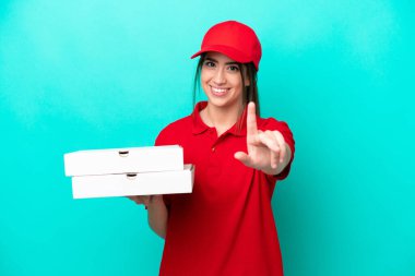 İş üniformalı bir pizzacı kadın mavi arka planda izole edilmiş pizza kutularını topluyor ve parmağını kaldırıyor.