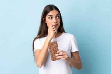 Mavi arka planda izole edilmiş beyaz bir kadın çikolata tableti alıyor ve kuşkuları var.