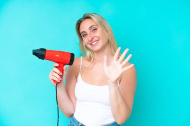 Beyaz bir kadın elinde saç kurutma makinesiyle mavi arka planda izole edilmiş bir şekilde elinde mutlu bir ifadeyle selam veriyor.