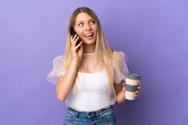 Mor arka planda izole edilmiş genç sarışın kadın elinde kahve ve bir cep telefonu tutuyor.