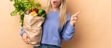 Bir çanta dolusu sebzeyi soyutlanmış arka planda tutan genç sarışın kadın şaşırmış ve parmağını kenarı işaret ediyor.
