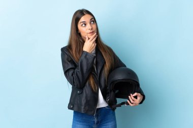 Mavi arka planda motosiklet kaskı tutan beyaz bir kadın gülümserken yukarı bakıyor.