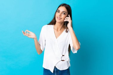 Mavi arka planda izole edilmiş genç beyaz bir kadın cep telefonuyla sohbet ediyor.