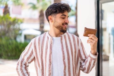 Yakışıklı bir Arap açık havada mutlu bir ifadeyle cüzdan tutuyor.