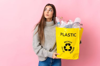 Young caucasian woman holding a bag full of plastic bottles to recycle isolated on pink background and looking up