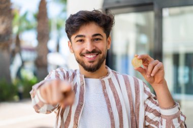 Açık havada Bitcoin tutan yakışıklı bir Arap, kendine güvenen bir ifadeyle seni işaret ediyor.