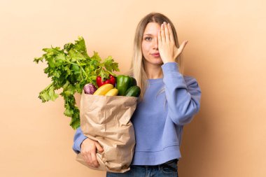 Elinde sebze dolu bir çanta tutan genç sarışın bir kadın izole edilmiş arka planda bir gözü elle kapatıyor.