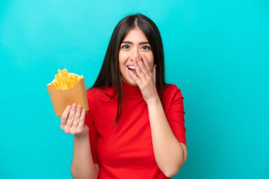 Fransız kızartması yakalayan beyaz bir kadın mavi arka planda izole edilmiş, şaşırmış ve şok olmuş bir yüz ifadesi var.