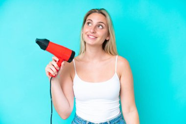 Mavi arka planda saç kurutma makinesi tutan beyaz bir kadın gülümserken yukarı bakıyor.