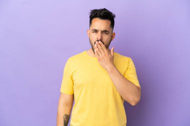 Mor arka planda izole edilmiş genç beyaz adam esniyor ve geniş bir ağzı eliyle kapatıyor.