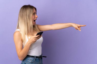 Mor arka planda izole edilmiş cep telefonu kullanan genç sarışın kadın parmağıyla kenarı işaret ediyor.