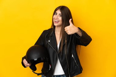 Sarı arka planda izole edilmiş motosiklet kaskı tutan beyaz bir kadın var. Başparmakları havada çünkü iyi bir şey oldu.