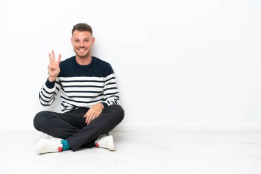 Young man sitting on the floor isolated on white background happy and counting three with fingers