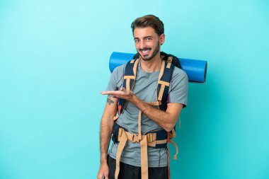 Mavi arka planda izole edilmiş büyük sırt çantalı genç dağcı bir fikir sunuyor.