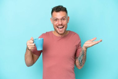 Kafkasyalı genç bir adam elinde kahve fincanıyla mavi arka planda izole edilmiş, şok olmuş bir yüz ifadesi var.