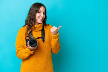 Mavi arka planda izole edilmiş genç fotoğrafçı kadın bir ürünü sunmak için kenarı işaret ediyor.