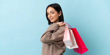 Mavi arka planda izole edilmiş genç beyaz bir kadın alışveriş torbaları tutuyor ve gülümsüyor.