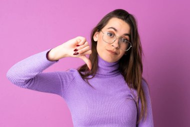 İzole edilmiş mor arka planda genç bir kadın baş parmağını negatif ifade ile aşağı gösteriyor.
