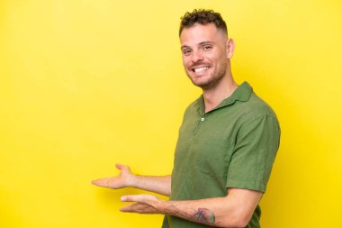 Sarı arka planda izole edilmiş beyaz tenli yakışıklı adam, davet edilmek için ellerini yana uzatıyor.