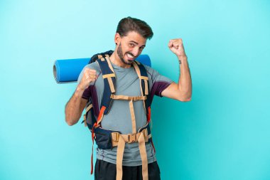 Mavi arka planda büyük bir sırt çantası olan genç dağcı zaferi kutluyor.