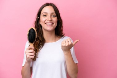 Pembe arka planda saç tarağı olan beyaz bir kadın bir ürünü sunmak için yan tarafı işaret ediyor.