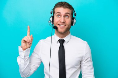 Telemarketer caucasian man working with a headset isolated on blue background pointing up and surprised