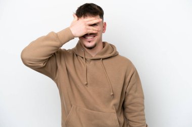 Beyaz arka planda izole edilmiş beyaz tenli yakışıklı bir adam. Gözleri elleriyle kaplıyor ve gülümsüyor.