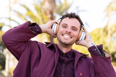 Genç beyaz bir adam açık havada müzik dinliyor.