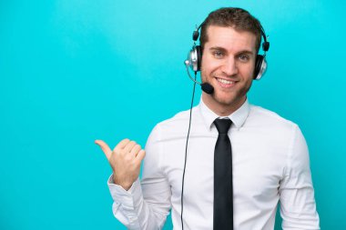 Tele pazarlamacı beyaz bir adam mavi arka planda izole edilmiş bir kulaklıkla çalışıyor. Yan tarafı göstererek bir ürün sunuyor.